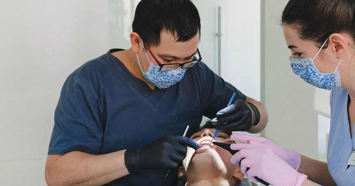 Two dental professionals in masks and gloves working together on a patient reclined in a chair in a bright modern clinic