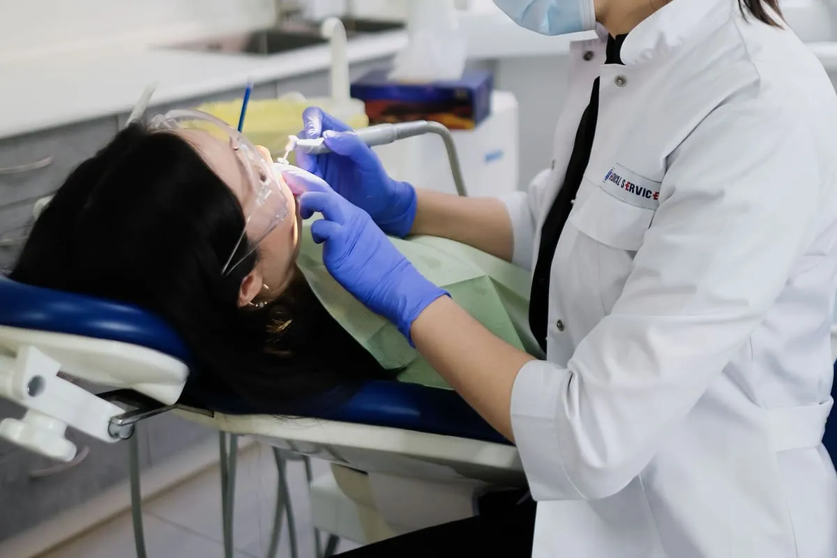 A clinician in a white coat leaning over a patient to clean their teeth, working in the close, static posture typical of chairside hygiene