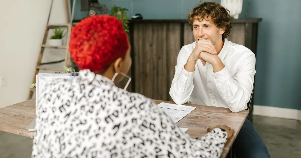 A man in a white shirt sits across a wooden table from a woman with short red hair, a laptop and a sheet of paper between them, in a casual office