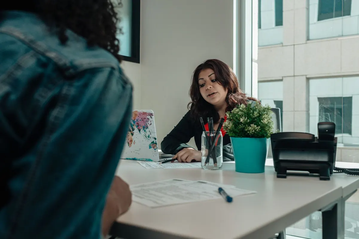 A woman with dark hair sits behind a white desk with a pen and papers while a person in a denim jacket sits facing her in the foreground