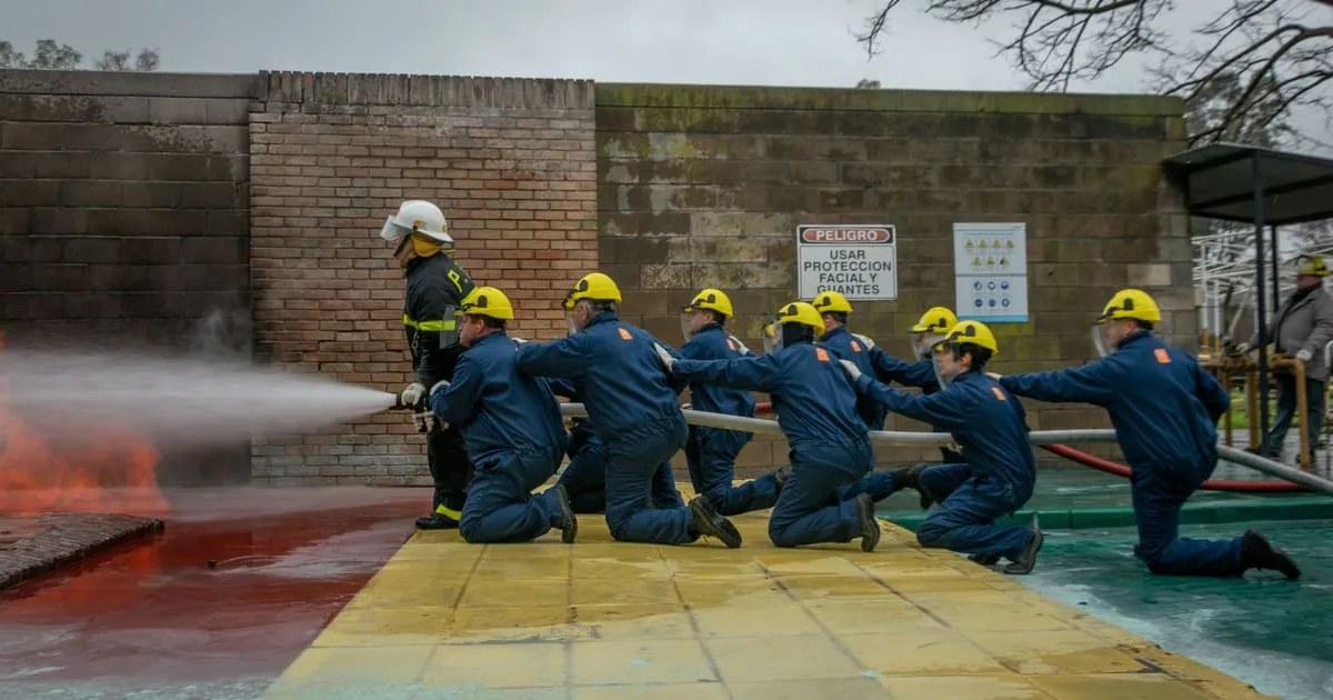 A line of firefighter trainees in yellow helmets and blue coveralls kneeling on a hose line during a live-fire training drill while an instructor directs the nozzle
