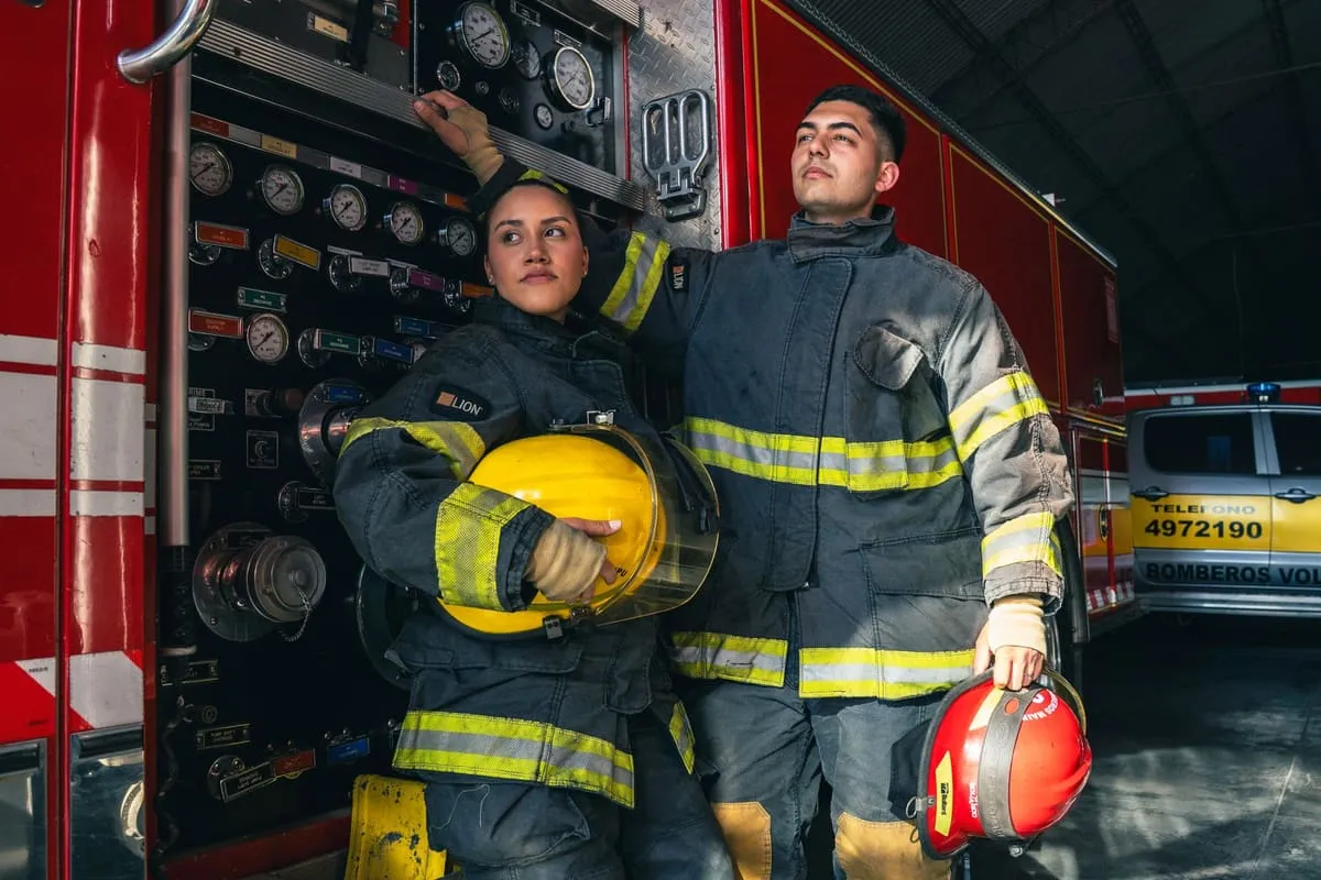 Two firefighters in turnout coats standing beside the pump panel of a red fire engine inside a station, one holding a yellow helmet and the other a red helmet
