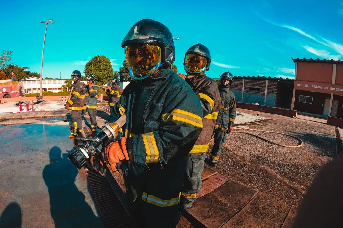 Firefighter recruits in full turnout gear and helmets with gold face shields standing in line on a training ground, the front recruit holding a hose nozzle