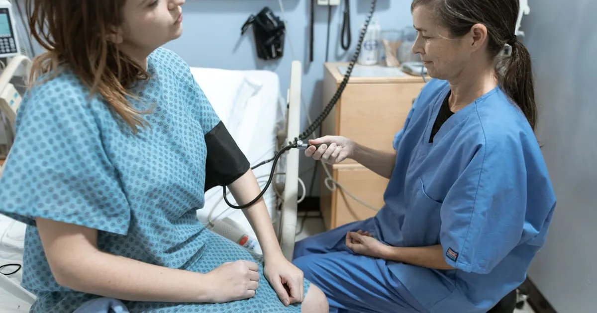 A healthcare worker in blue scrubs checking a patient's blood pressure with a manual cuff and stethoscope in a clinic exam room