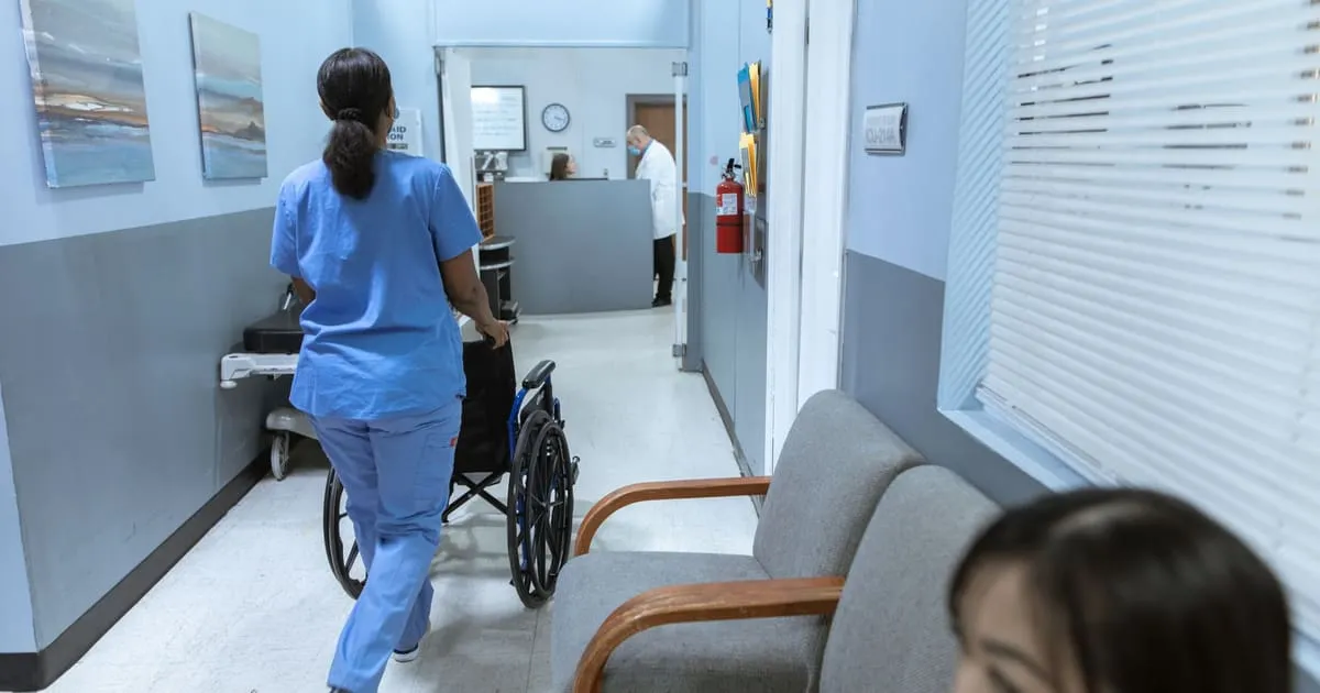 A nurse in blue scrubs pushes an empty wheelchair down a bright hospital hallway toward a reception desk