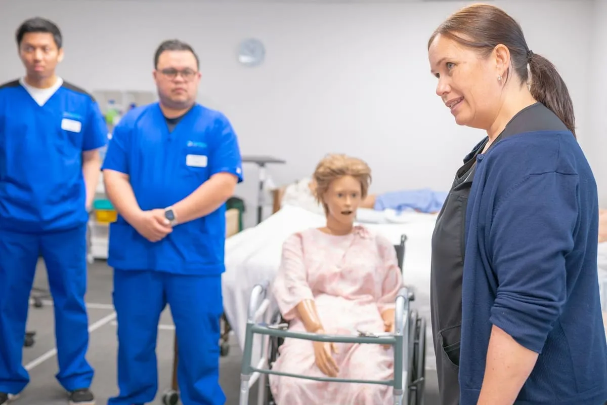 An adult nursing instructor in a navy cardigan speaks to two students in blue scrubs beside a medical mannequin seated in a wheelchair in a clinical skills lab