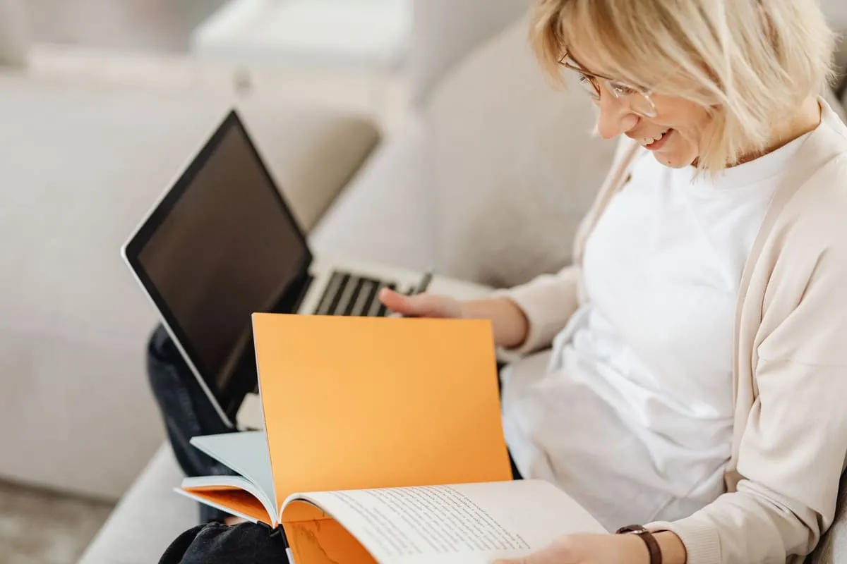 A woman with glasses sits on a couch smiling as she turns the pages of an open orange notebook with a laptop beside her, studying at home