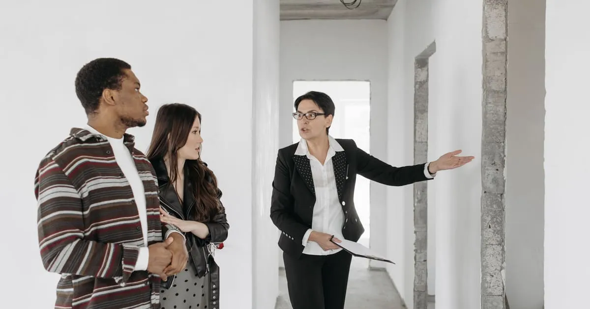 A real estate agent in a black blazer holding a clipboard points down the hallway of an unfinished house while a young couple listens