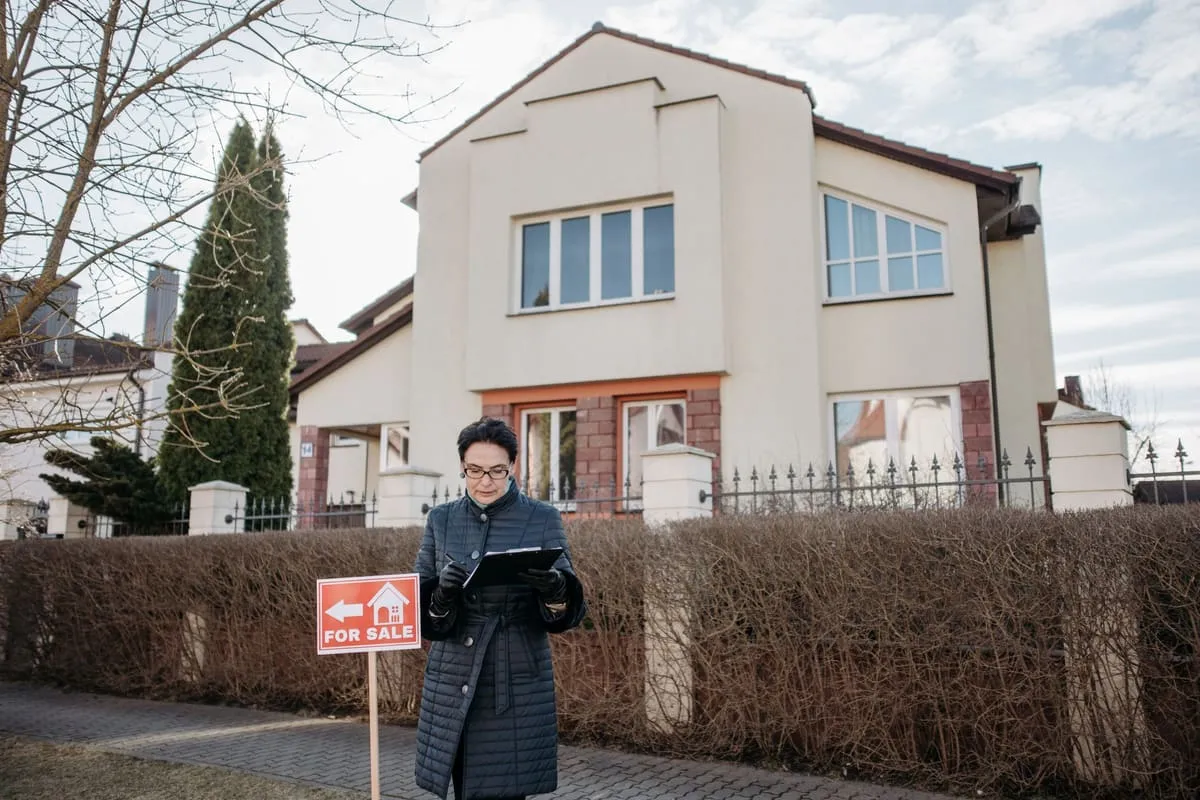 A middle-aged real estate agent in a long winter coat reviews notes on a clipboard on the sidewalk in front of a two-story house with a red For Sale sign beside her