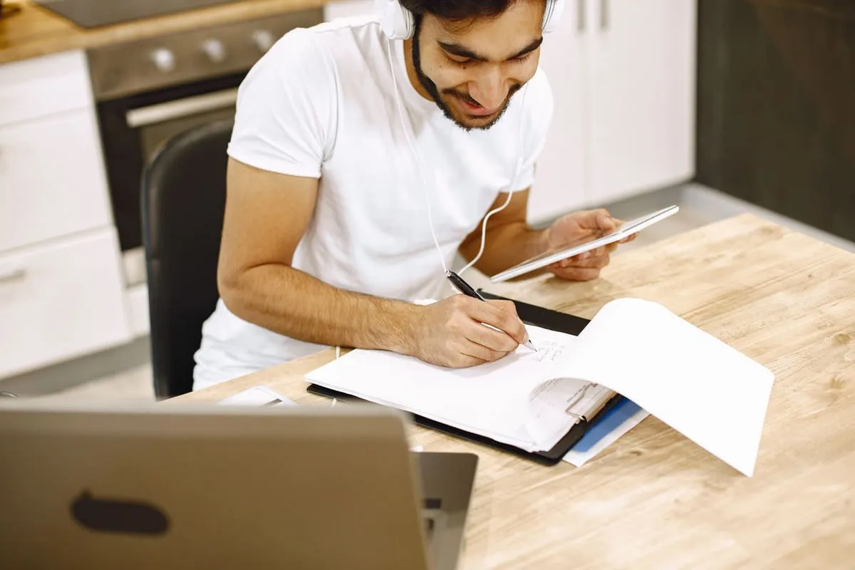 A man in a white T-shirt and headphones writes notes on printed course pages at a kitchen table, holding a tablet with a laptop open in front of him