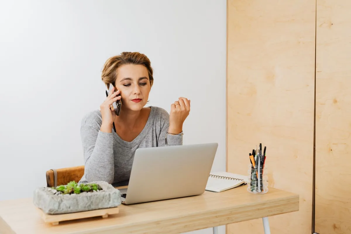 A person working from a home office desk on a laptop while taking a phone call, representing a home-based travel agent business